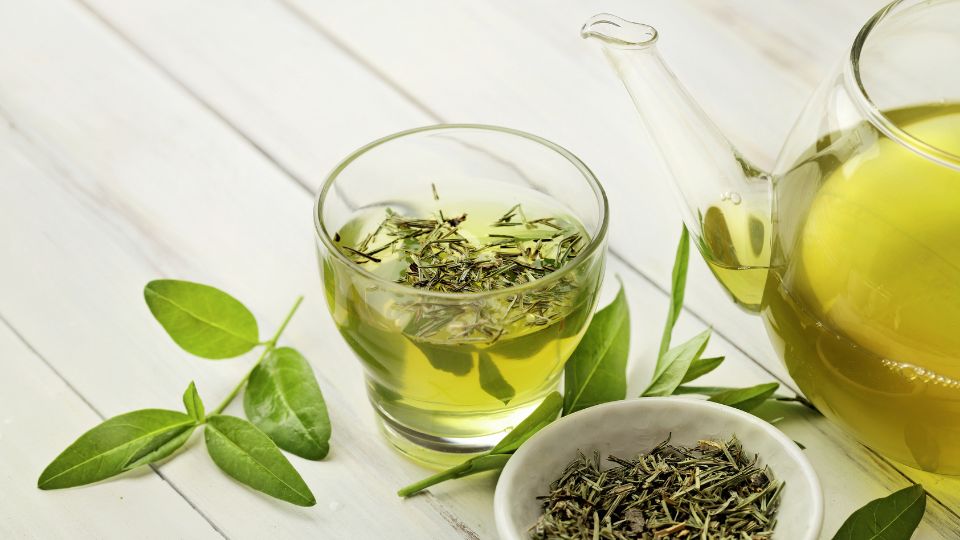 Friss zöld tea egy pohárban, mellette teáskanna és szárított levelek.